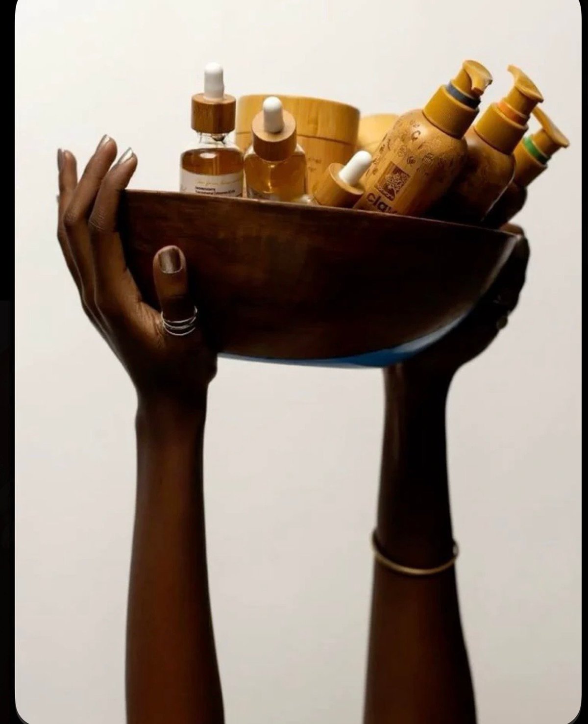Two hands holding a black bowl filled with golden beauty and haircare products against a white background