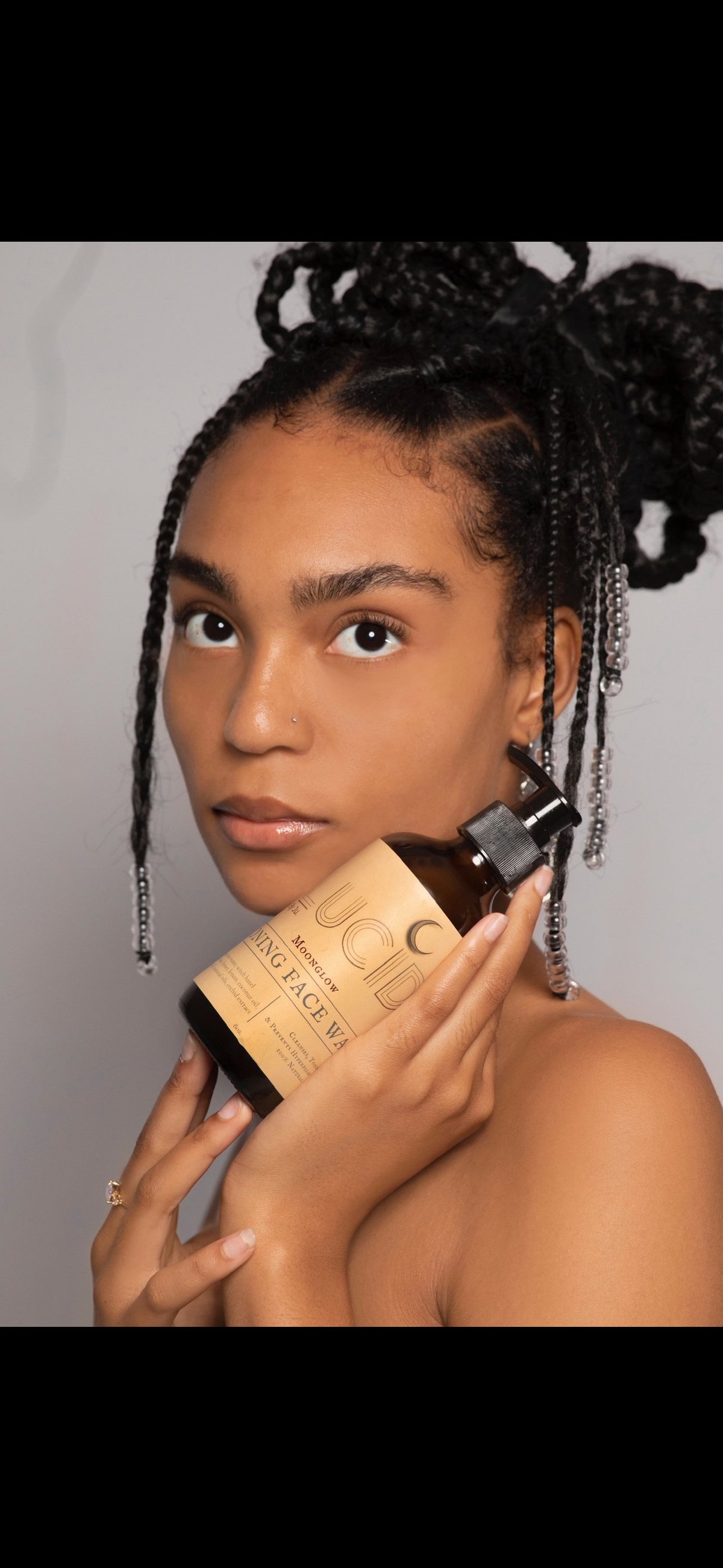 Young woman with braided hairstyle holding a foundation bottle near her face against a neutral gray background