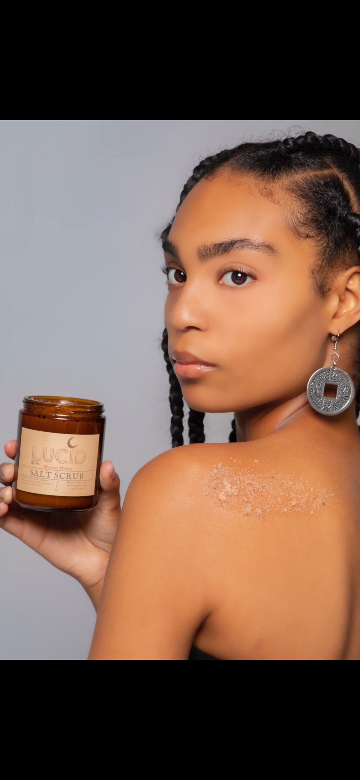Woman holding a jar of salt scrub product, with visible exfoliant on her shoulder against a neutral gray background