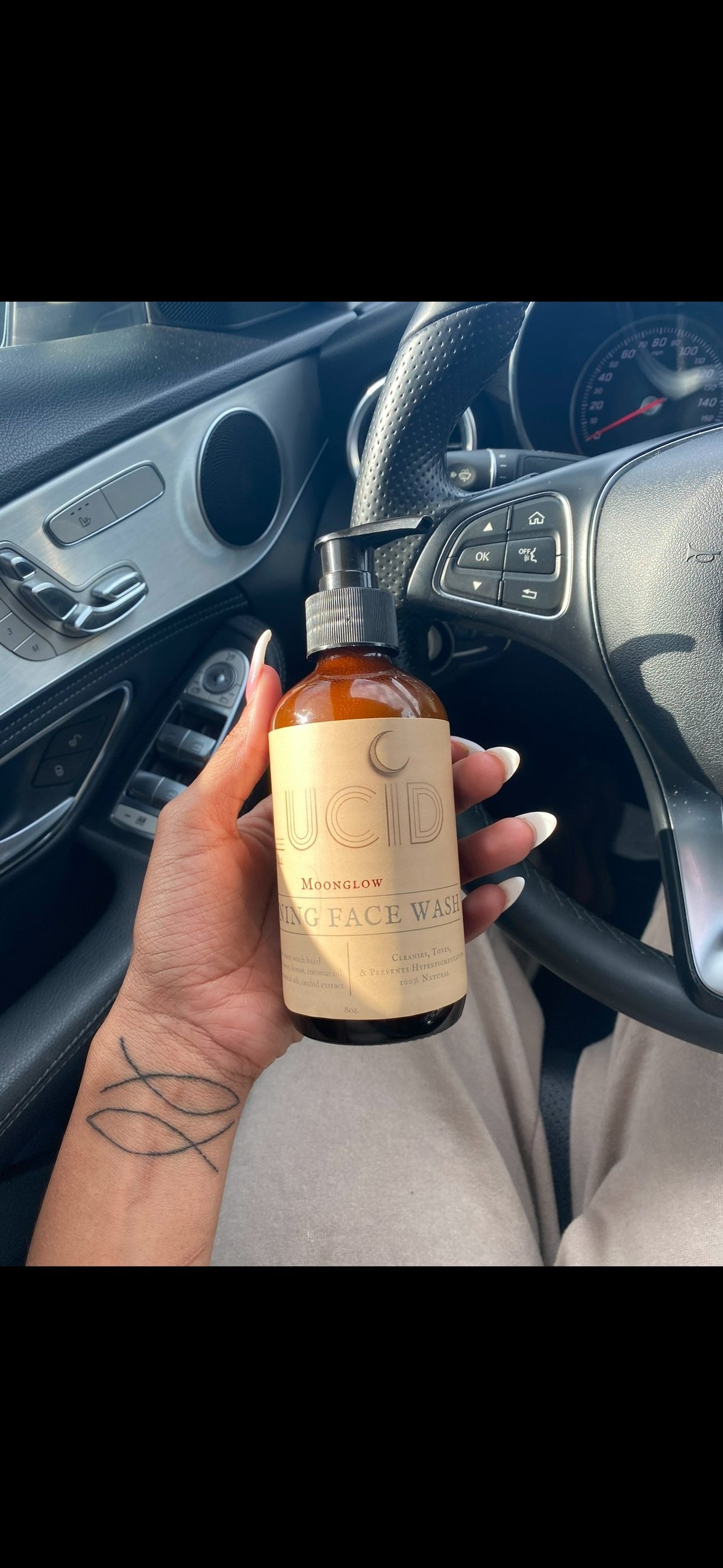 Person holding a bottle of ucd Moonglow face wash inside a car with steering wheel and dashboard visible